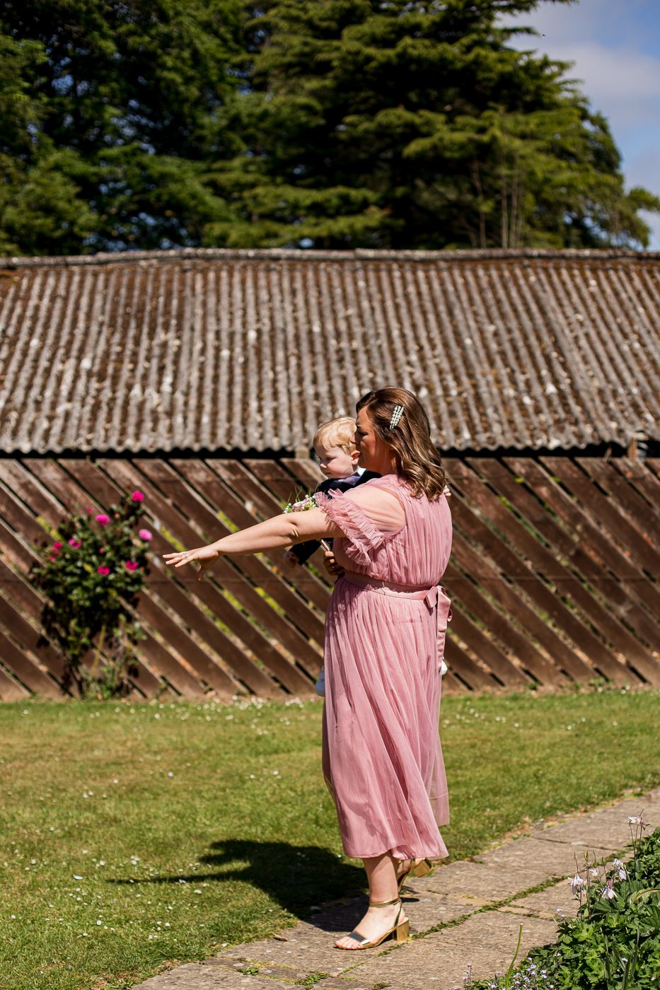 A pink-dressed figure poses playfully in a garden setting with a wooden fence backdrop.
