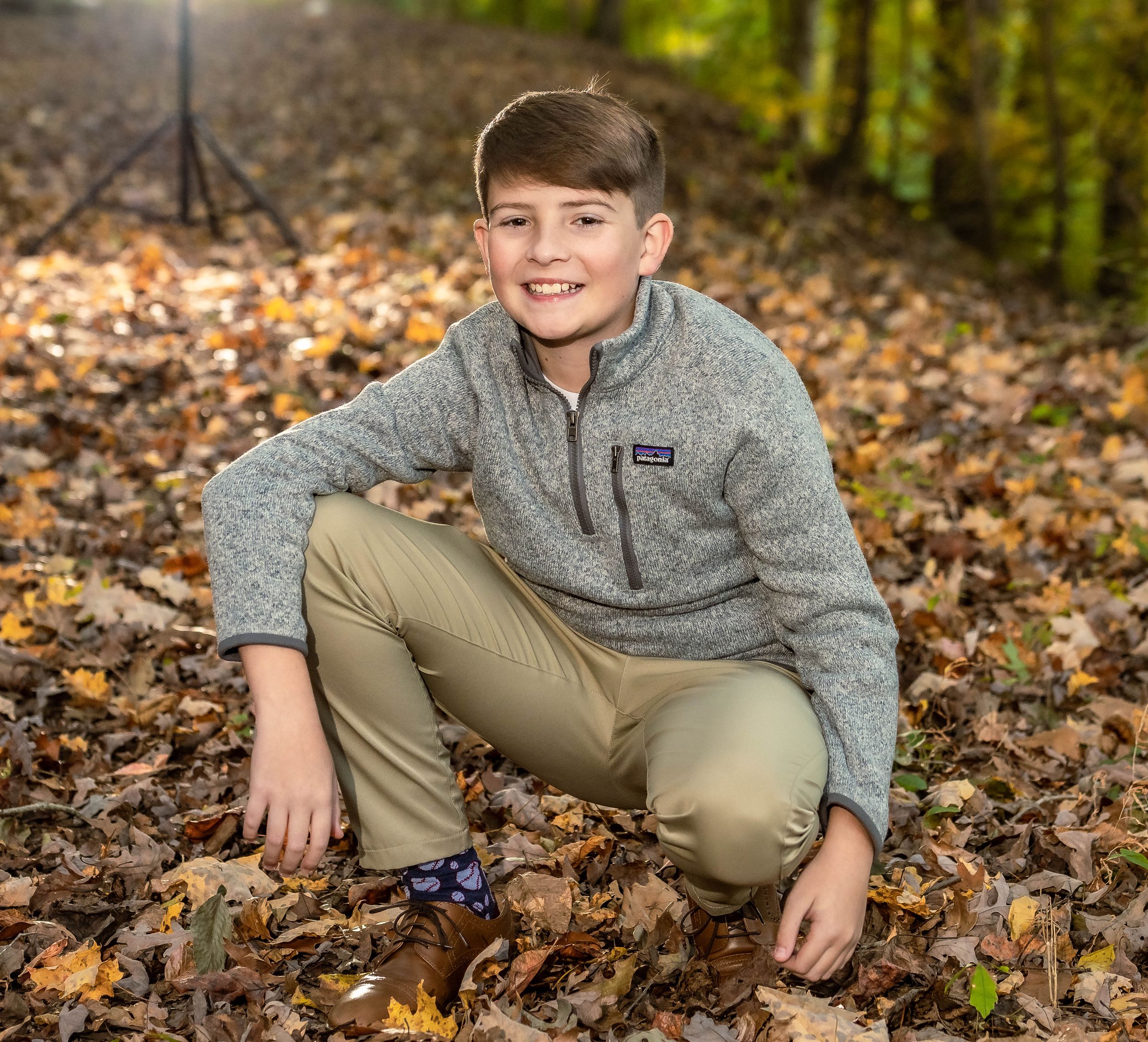A child wearing a gray fleece sweater and khaki pants lies in autumn leaves during an outdoor photo session.