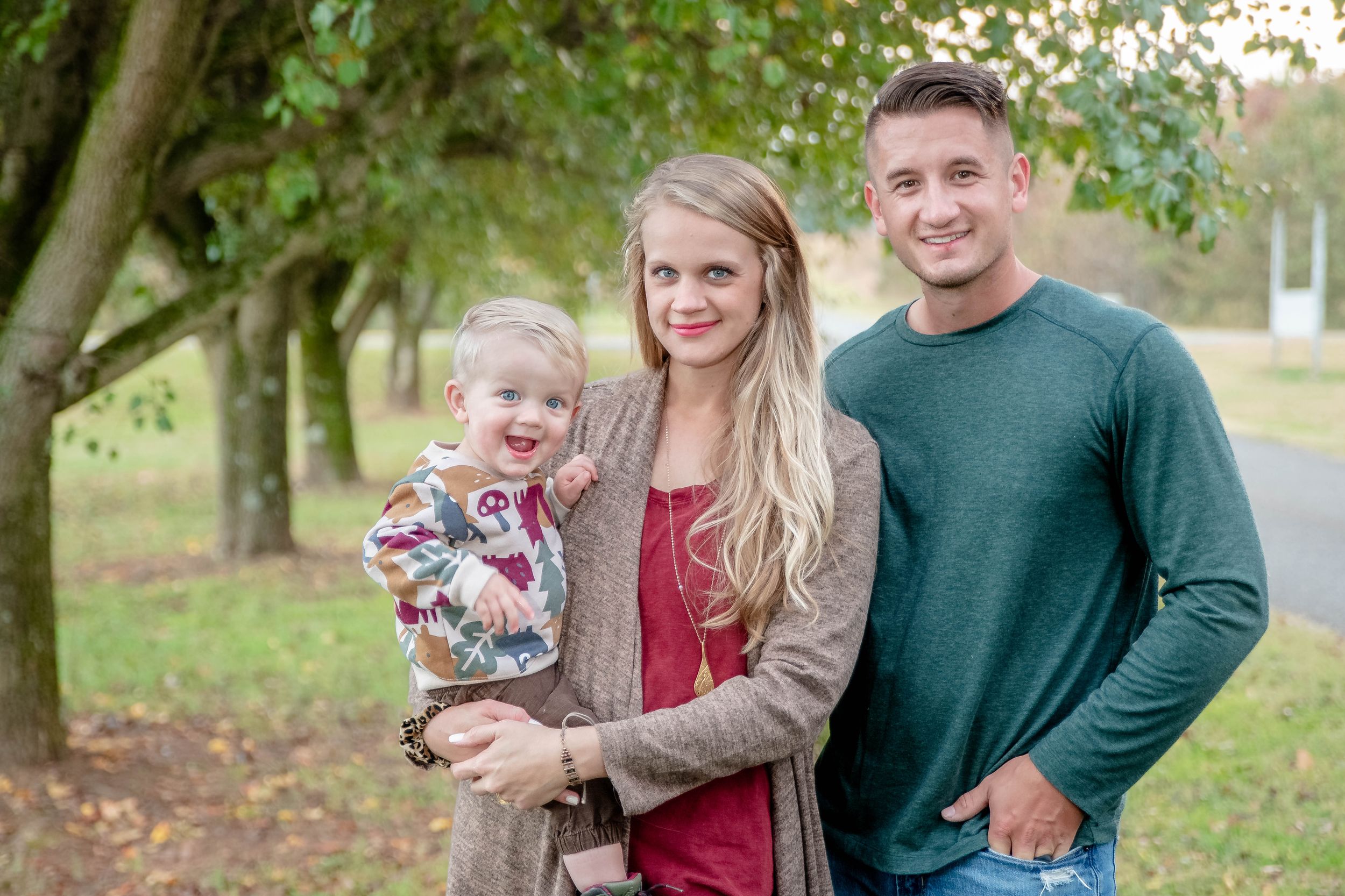 A family poses together outdoors in autumn, wearing casual sweaters against a backdrop of trees with fallen leaves.