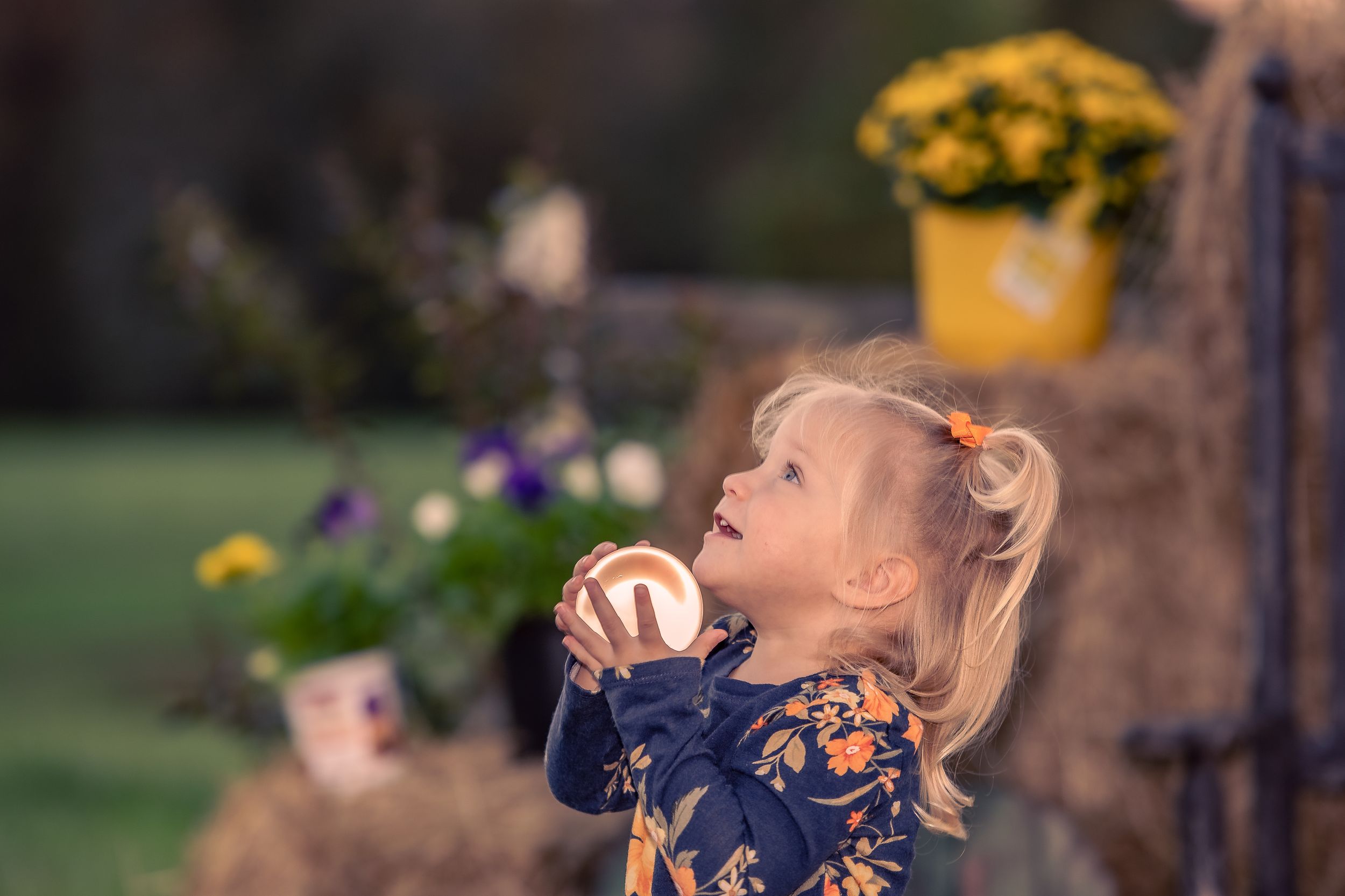 Young child in navy dress blowing bubbles outdoors near yellow flowers.
