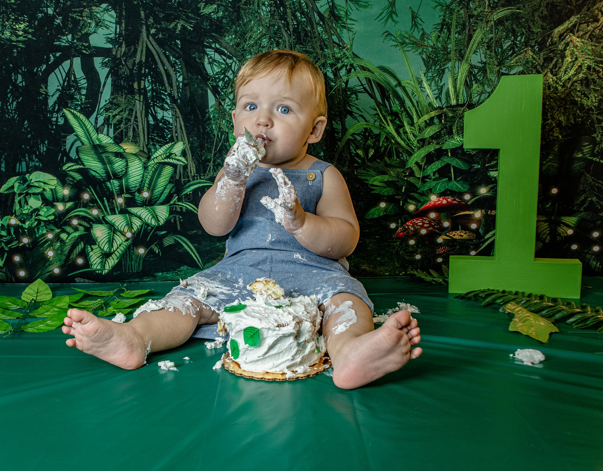 A baby sits on a green table wearing a white outfit during a cake smash photo session with jungle-themed backdrop.