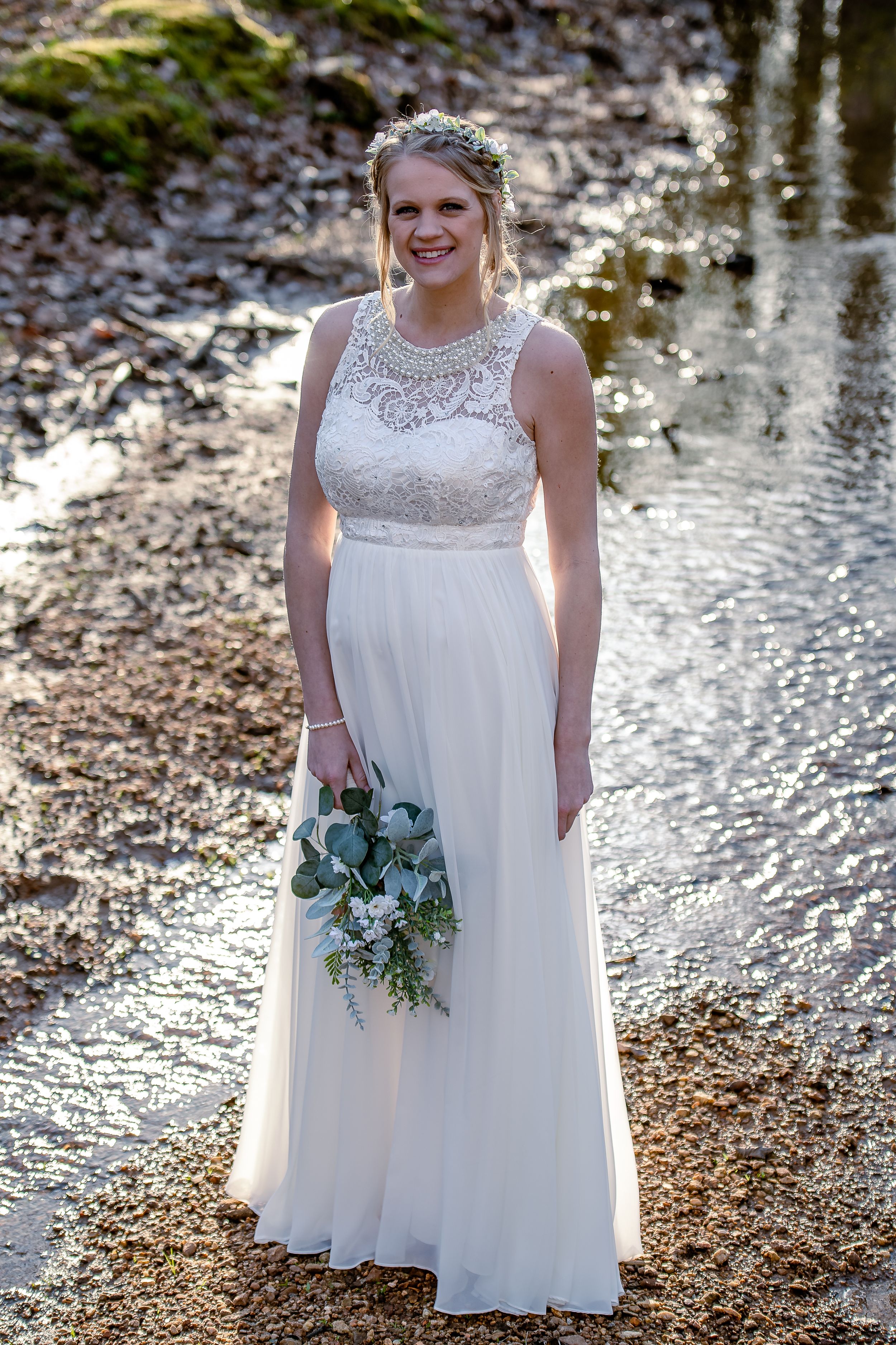 Bride in a flowing white lace-top dress poses by a reflective waterfront holding a blue-green floral bouquet.