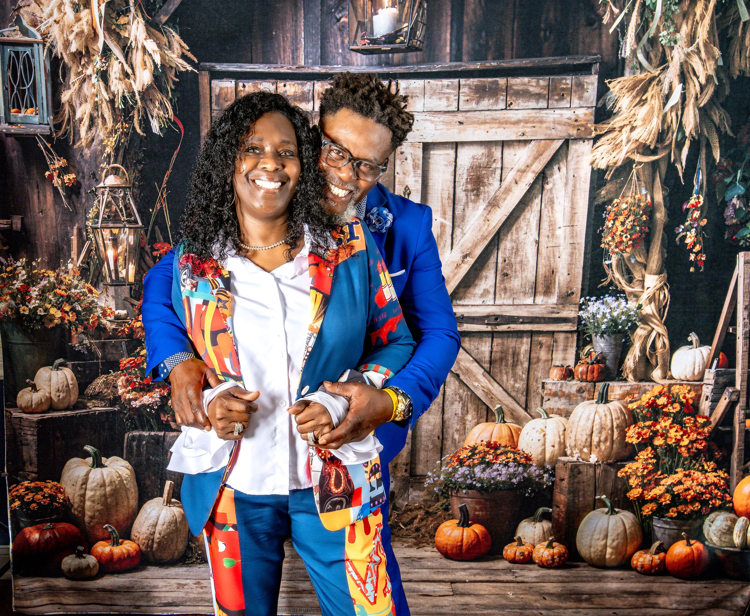 A couple in autumn attire poses together at a rustic fall display with pumpkins and dried corn stalks.