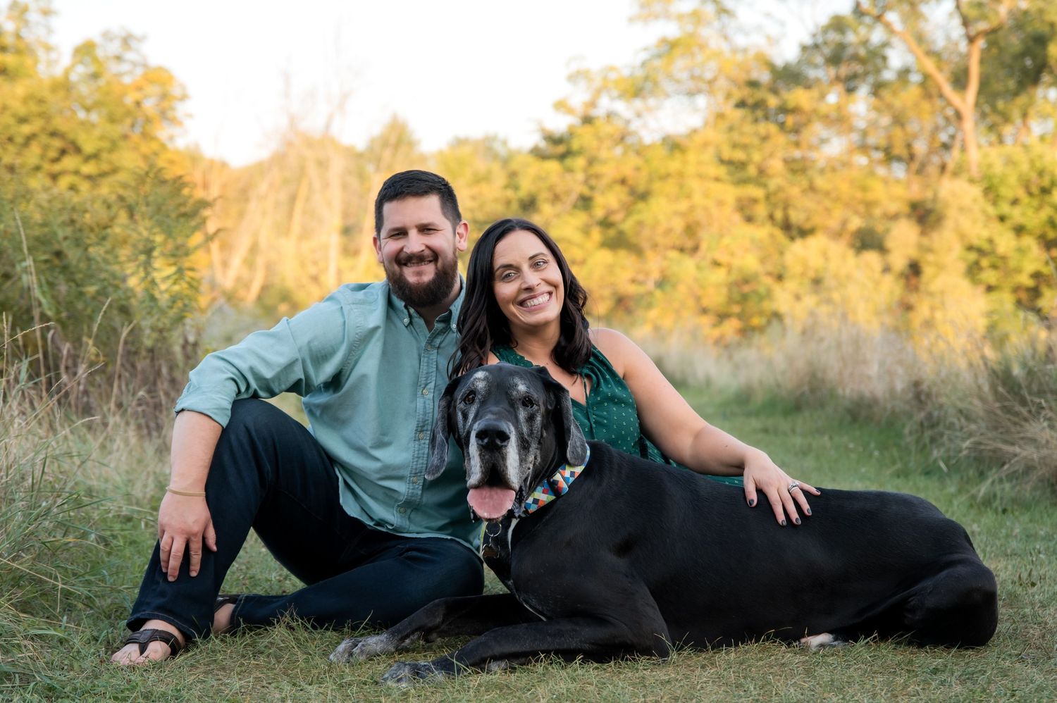 A couple and their black dog lay together in a grassy field during golden hour.