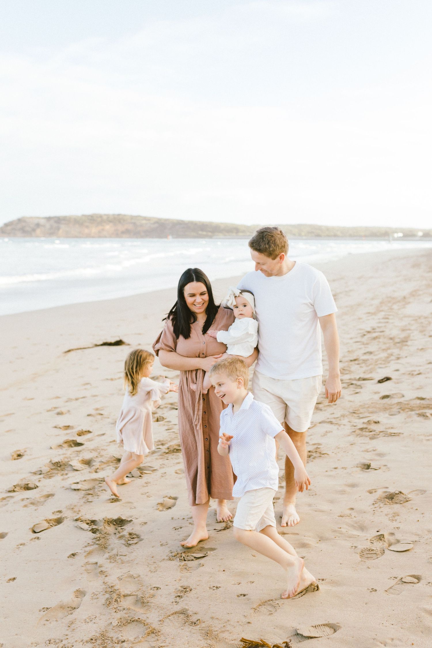 Family enjoying a playful moment together on a sandy beach wearing coordinated white and beige outfits.