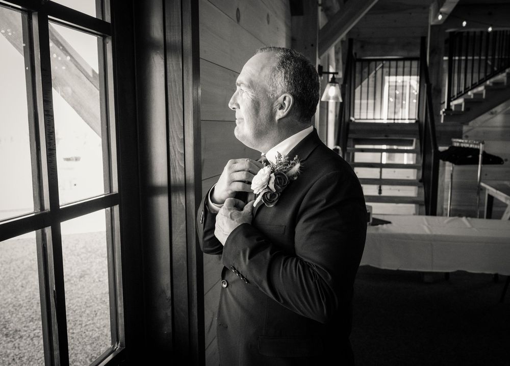 Black and white photo of a groom in a suit adjusting his tie while looking out a window in contemplative pose.