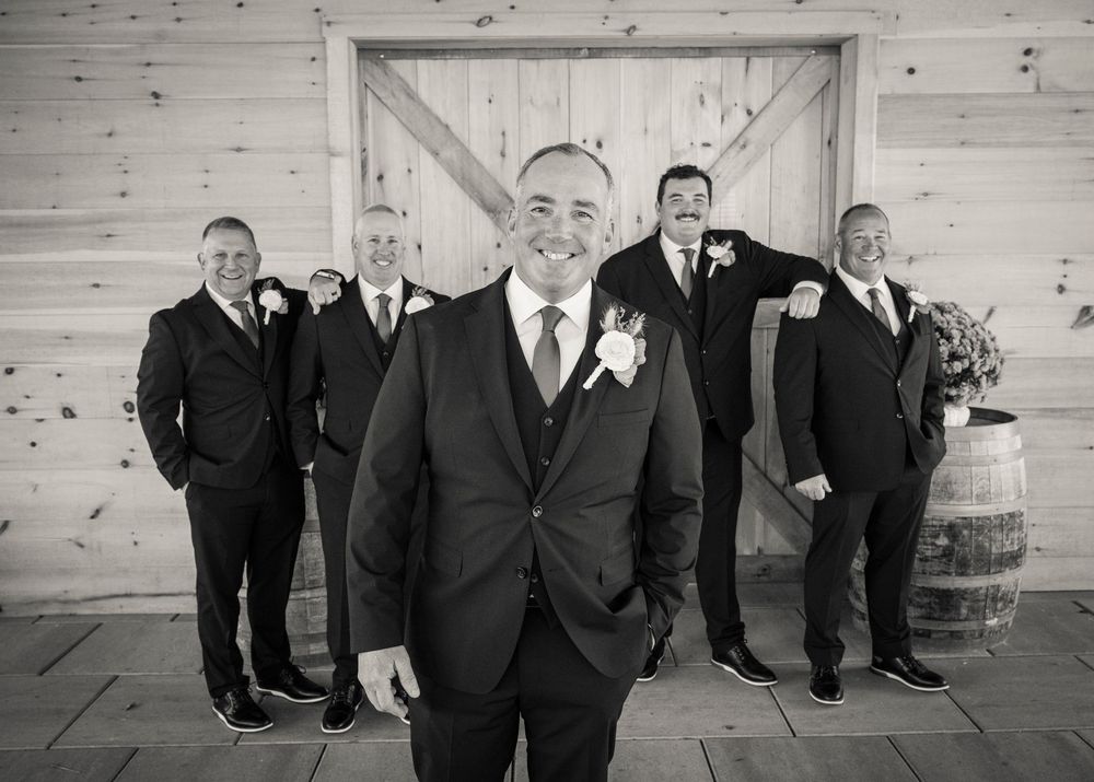 Groomsmen in black suits and ties pose together in front of a white wooden wall.