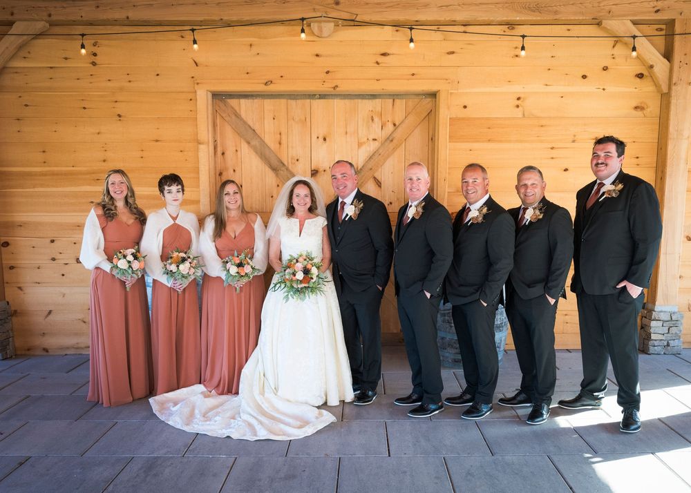 Wedding party members seated on wooden deck for formal group photos at rustic venue.