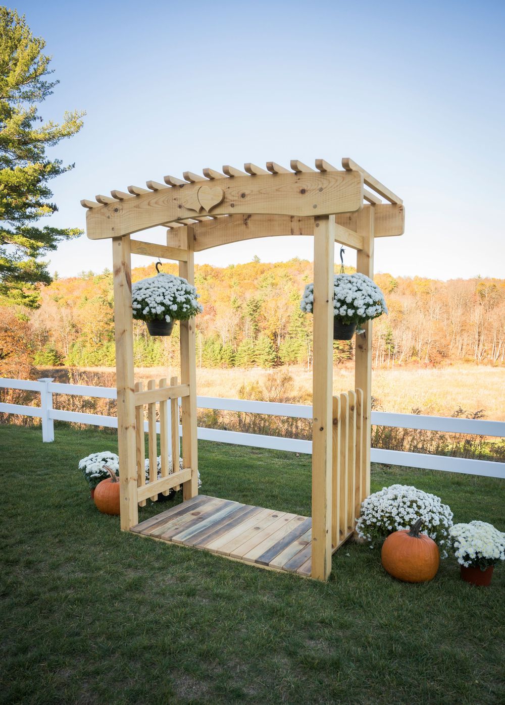 Wooden pergola decorated with white flowers and pumpkins stands on a grassy lawn with autumn foliage in the background.