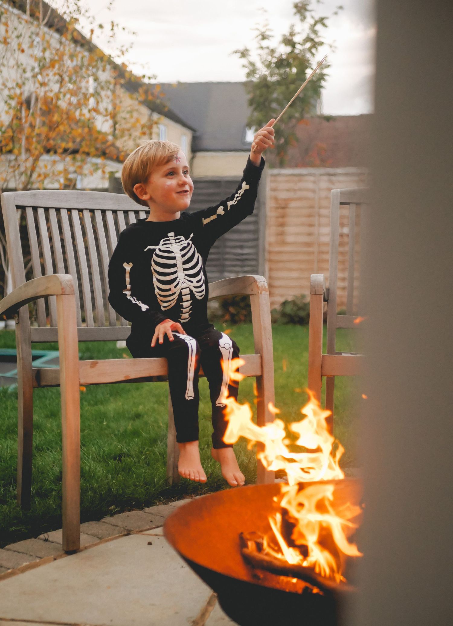 A child in a skeleton costume sits by a glowing orange fire pit on an autumn evening.