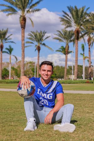 Baseball player in blue IMG jersey and gray pants sits on grass field with palm trees in background on sunny day.