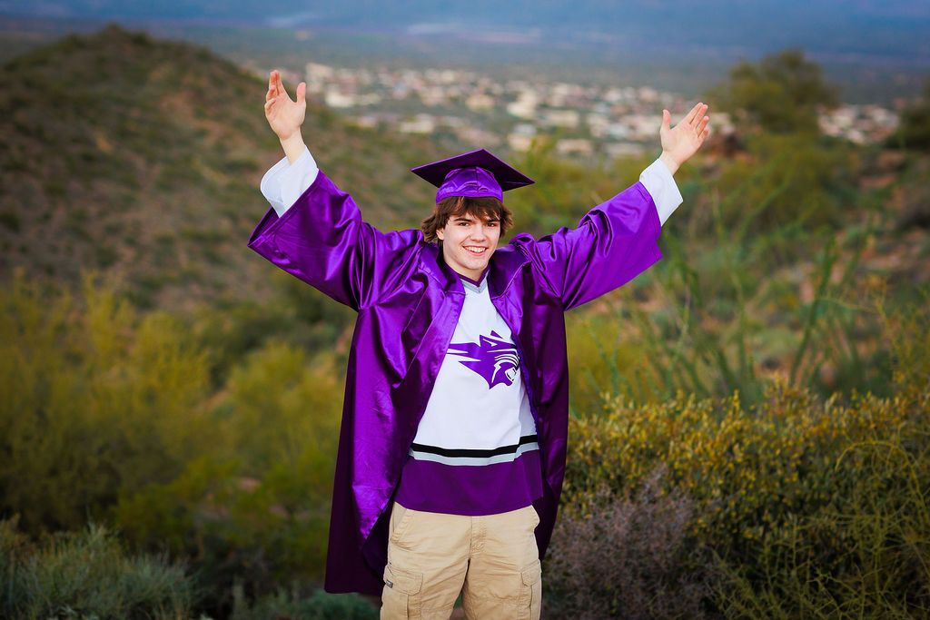Graduate in purple cap and gown celebrates with raised arms on hilltop overlooking city view.