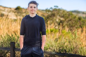 A person in a black t-shirt stands outdoors against a scenic natural landscape with golden grass and blue sky.