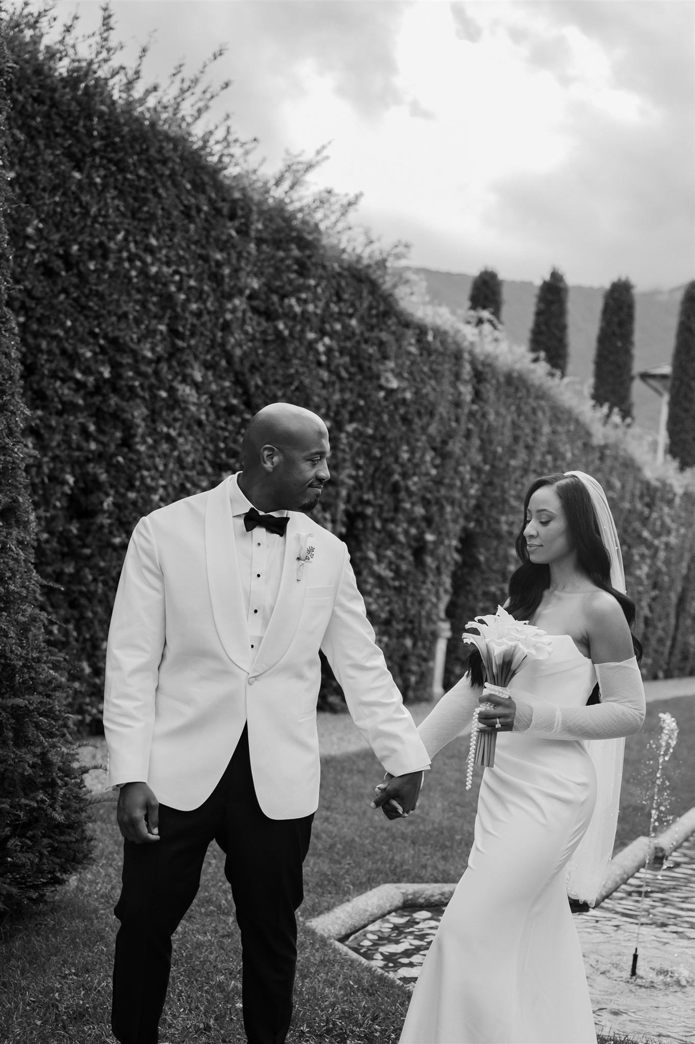 A couple in wedding attire dances playfully among tall hedges in a formal garden.