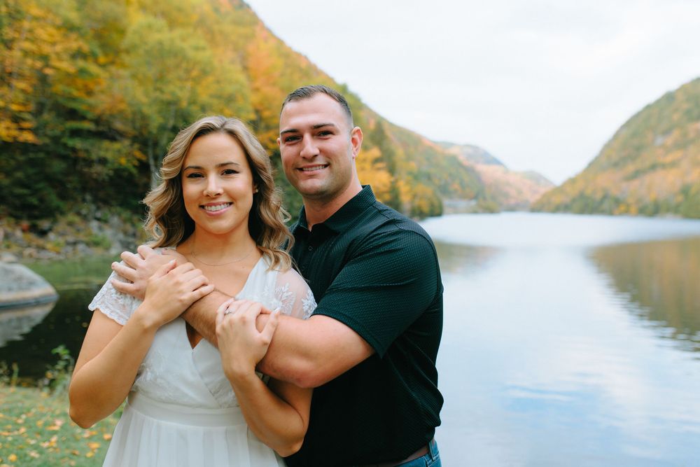 A couple embraces near a scenic lake surrounded by autumn foliage and mountains during a romantic photoshoot.