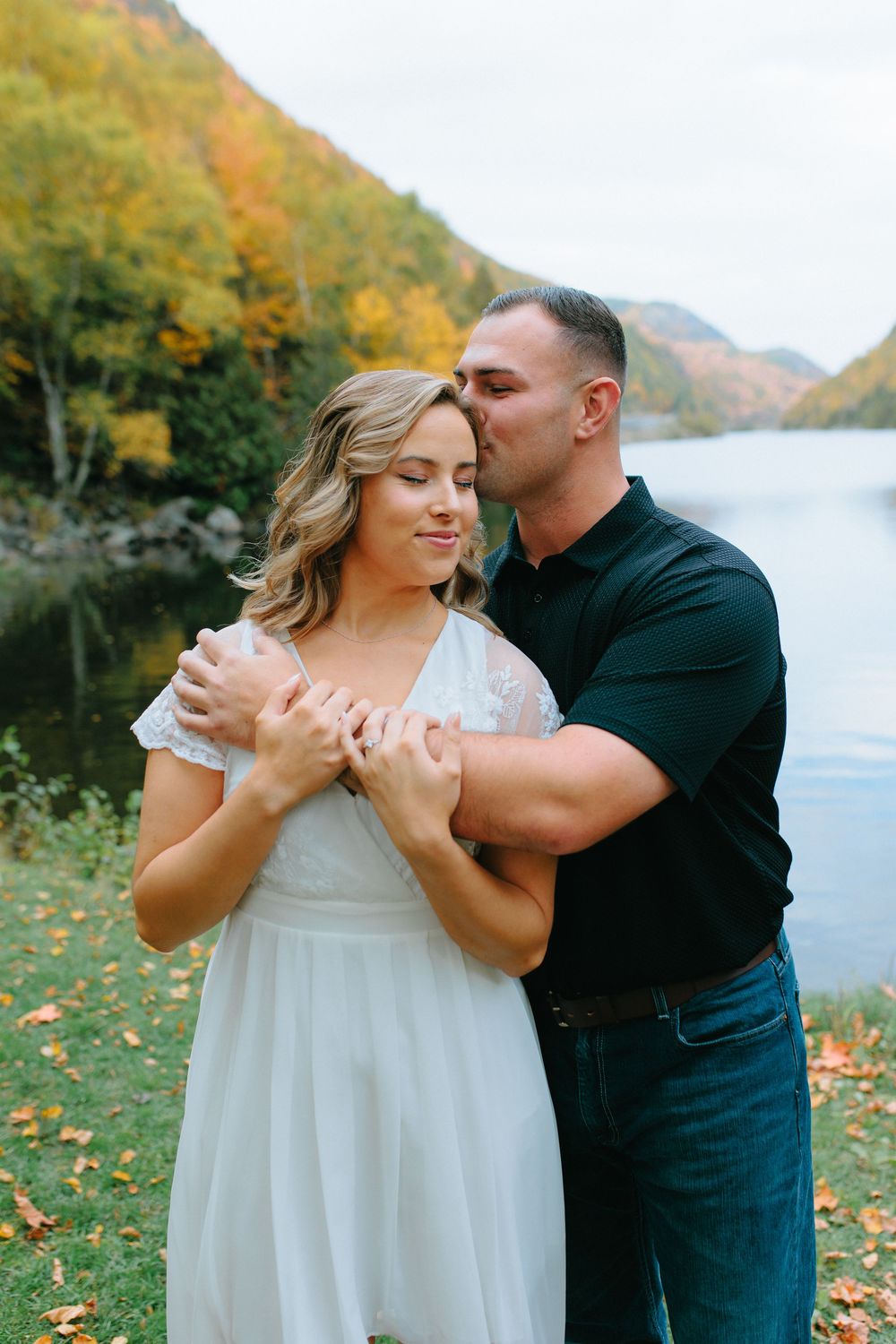 A couple embraces in an autumn setting by a lake with colorful fall foliage in the background.