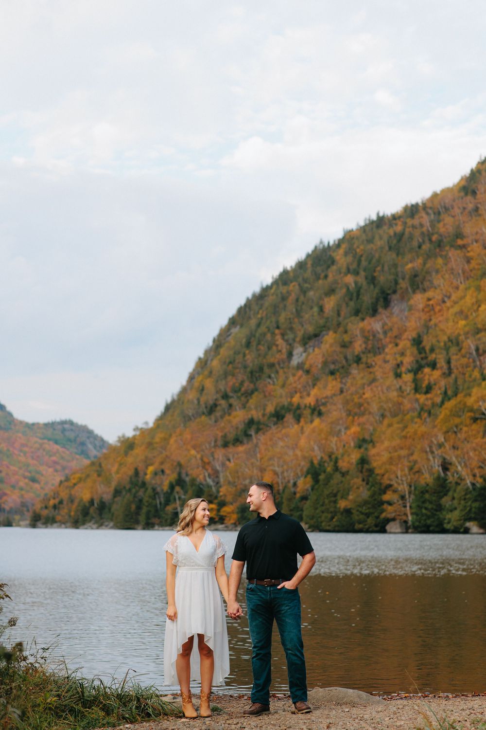 A couple stands together on a shoreline with stunning fall-colored mountains and a serene lake in the background.