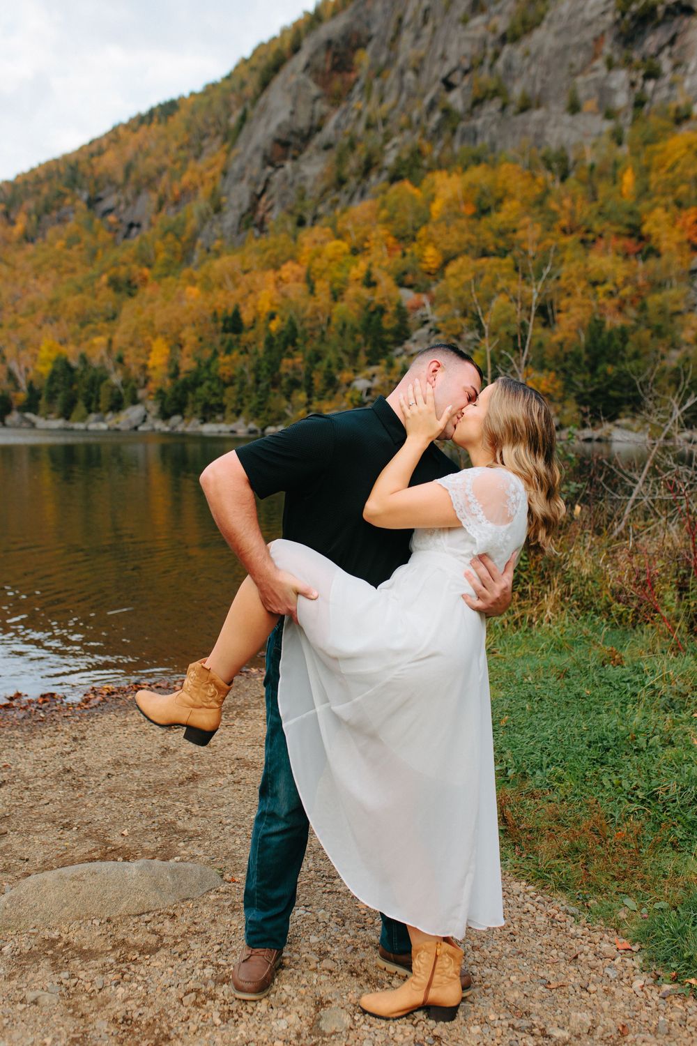 A romantic couple embraces by a lake surrounded by autumn foliage and mountain cliffs.
