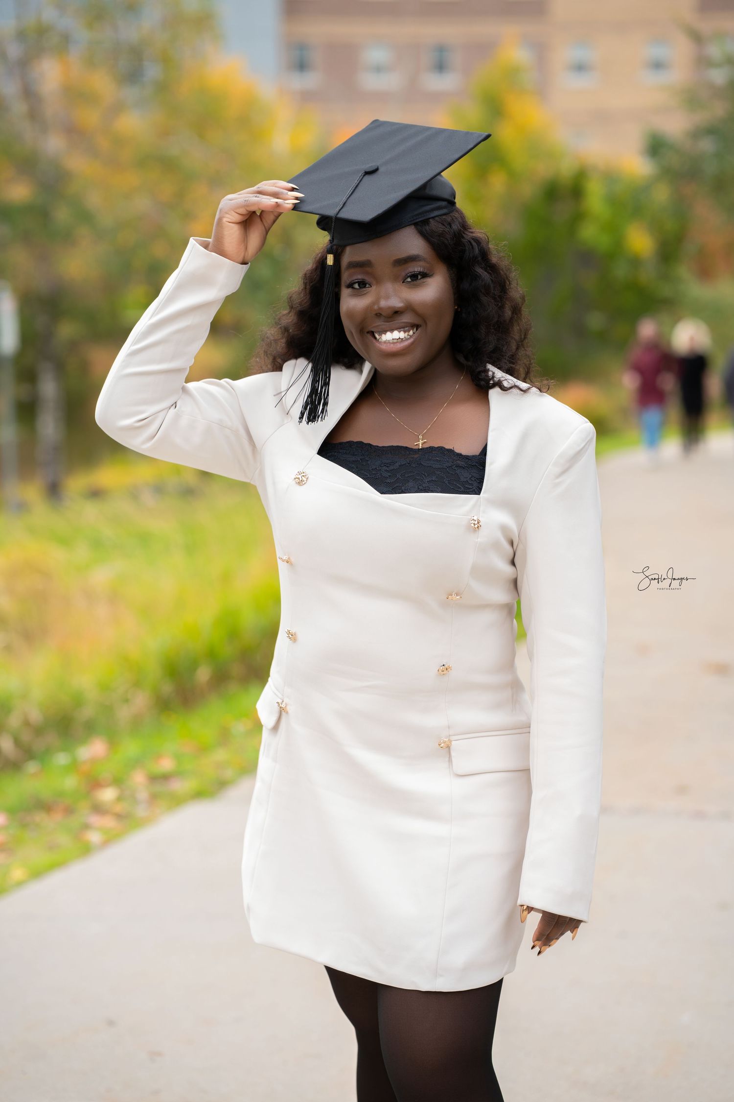 Graduate in white blazer and cap poses joyfully outdoors during graduation photoshoot on sunny autumn day.