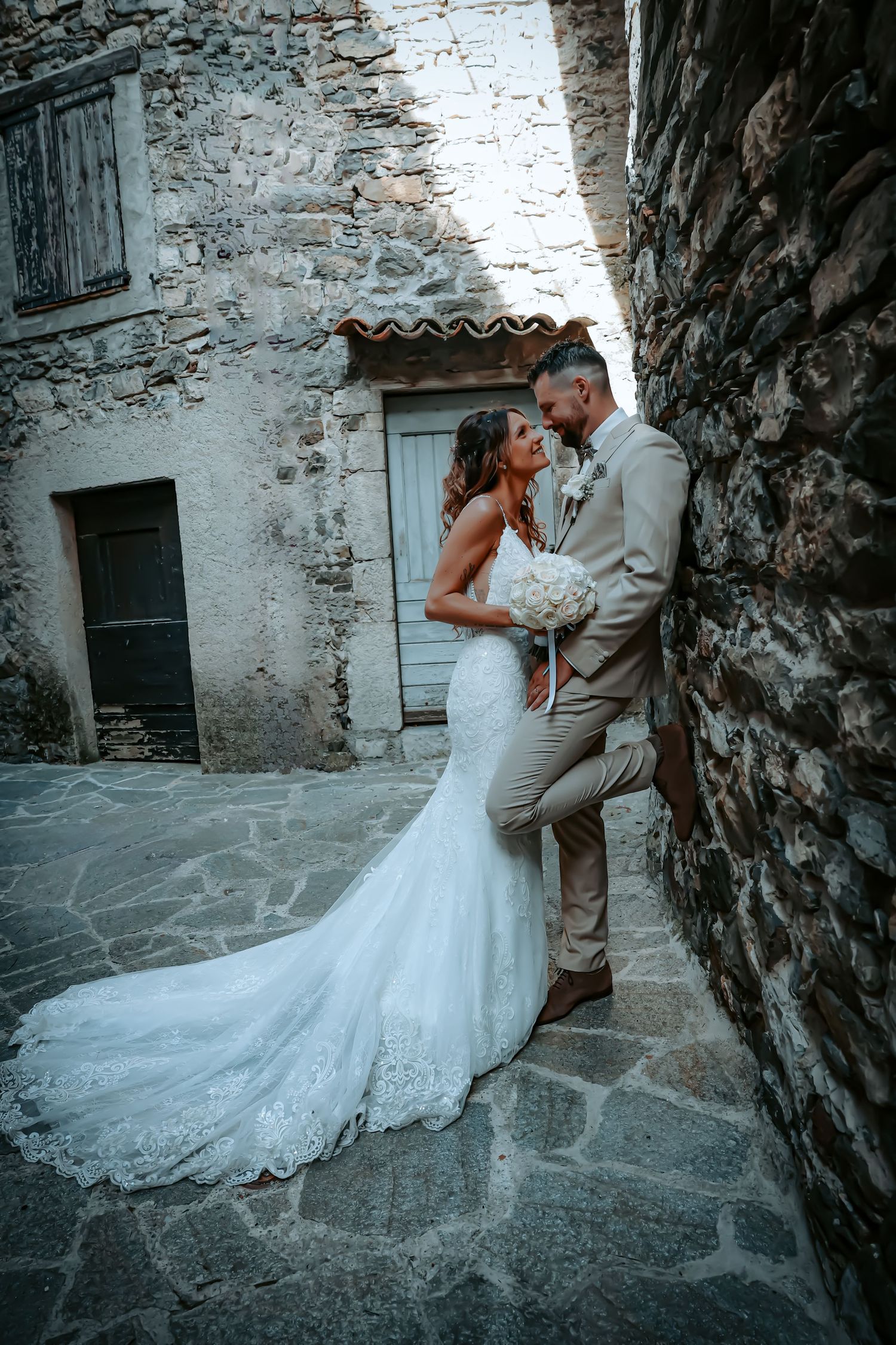 Bride in flowing white gown poses with groom along rustic stone walkway.