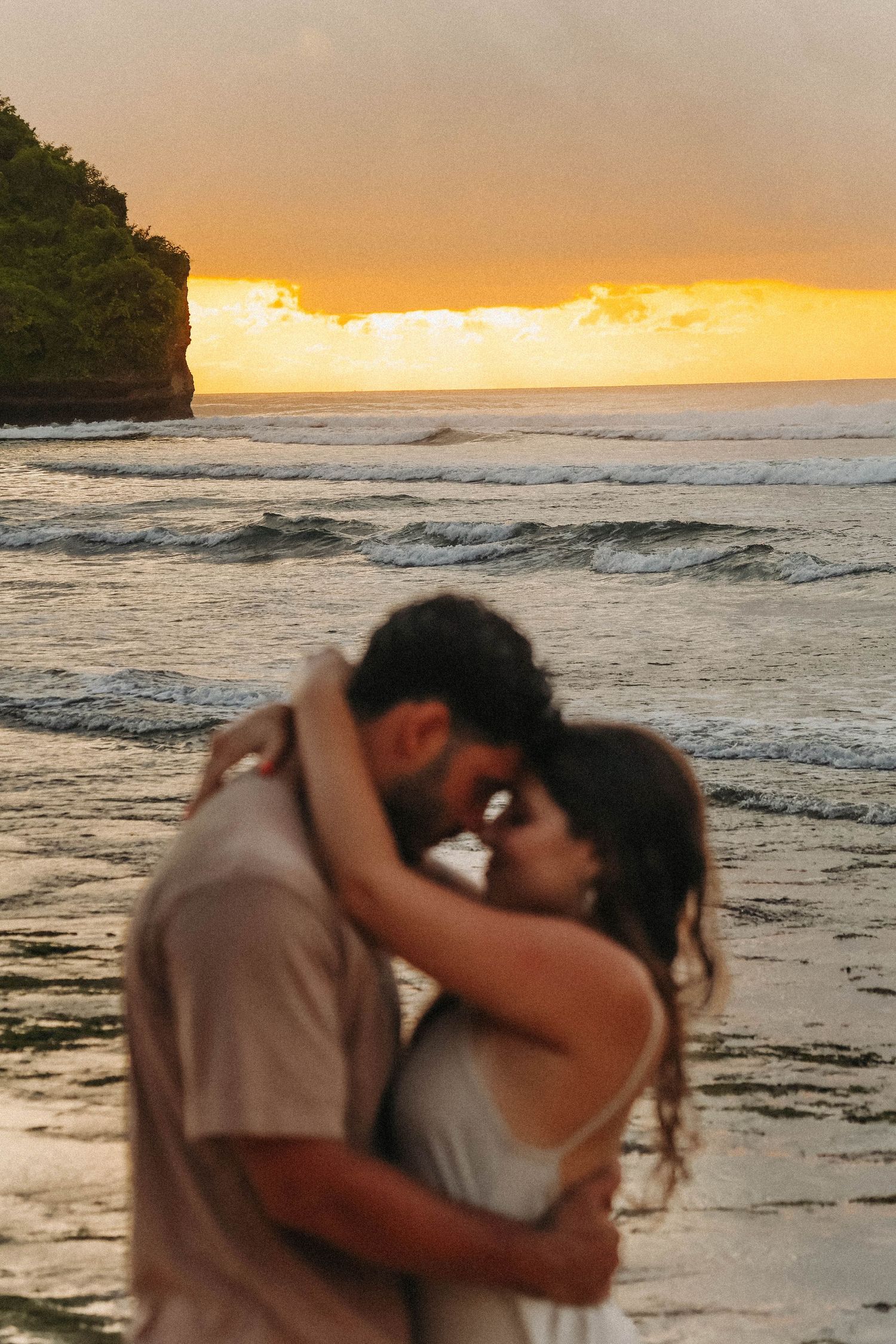 Couple sharing a kiss at golden sunset on tropical beach with ocean waves and rocky cliffs behind.