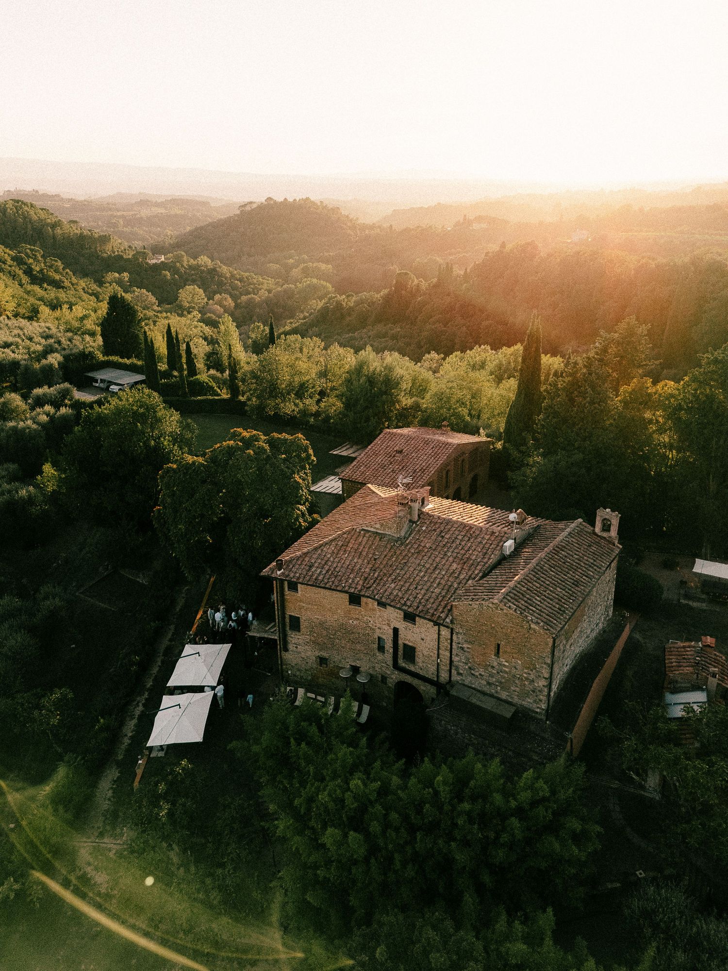 Aerial view of rustic Tuscan villa surrounded by cypress trees at golden sunset in Italian countryside.