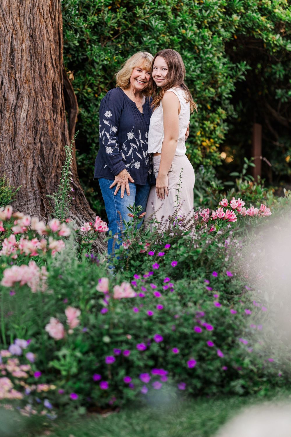 Pink and purple flowers bloom in a garden border with people sitting on a bench in soft focus behind them.