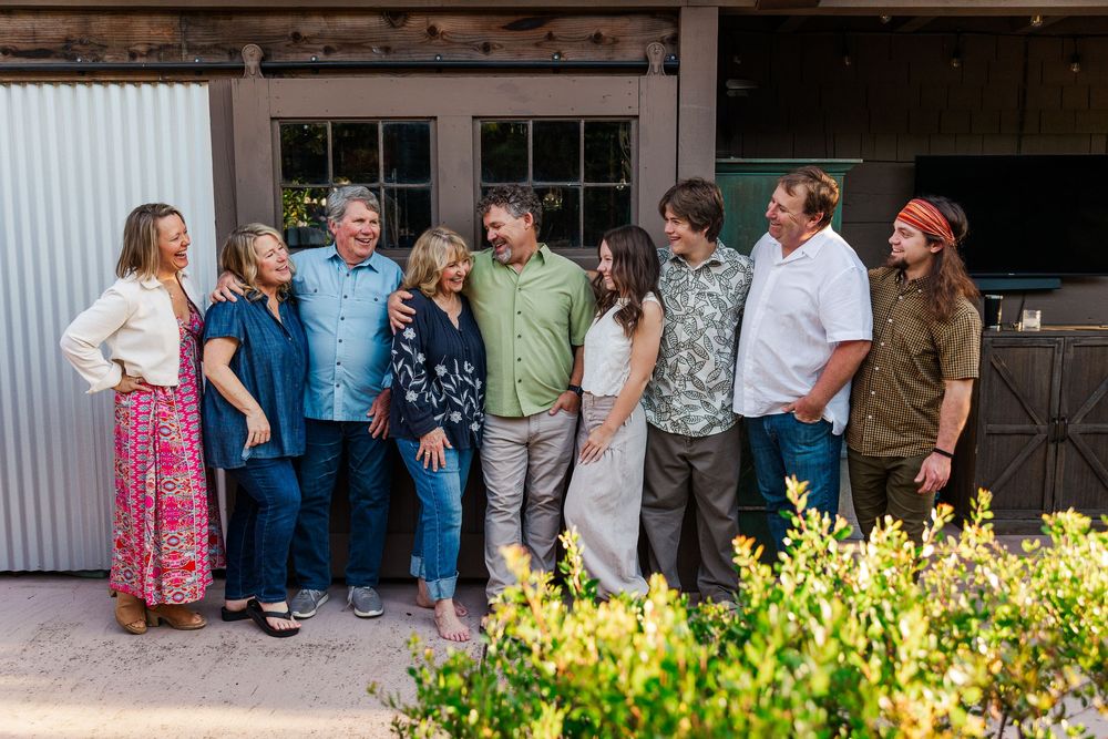 A group of people stand together smiling outside a rustic building with yellow flowers in the foreground.