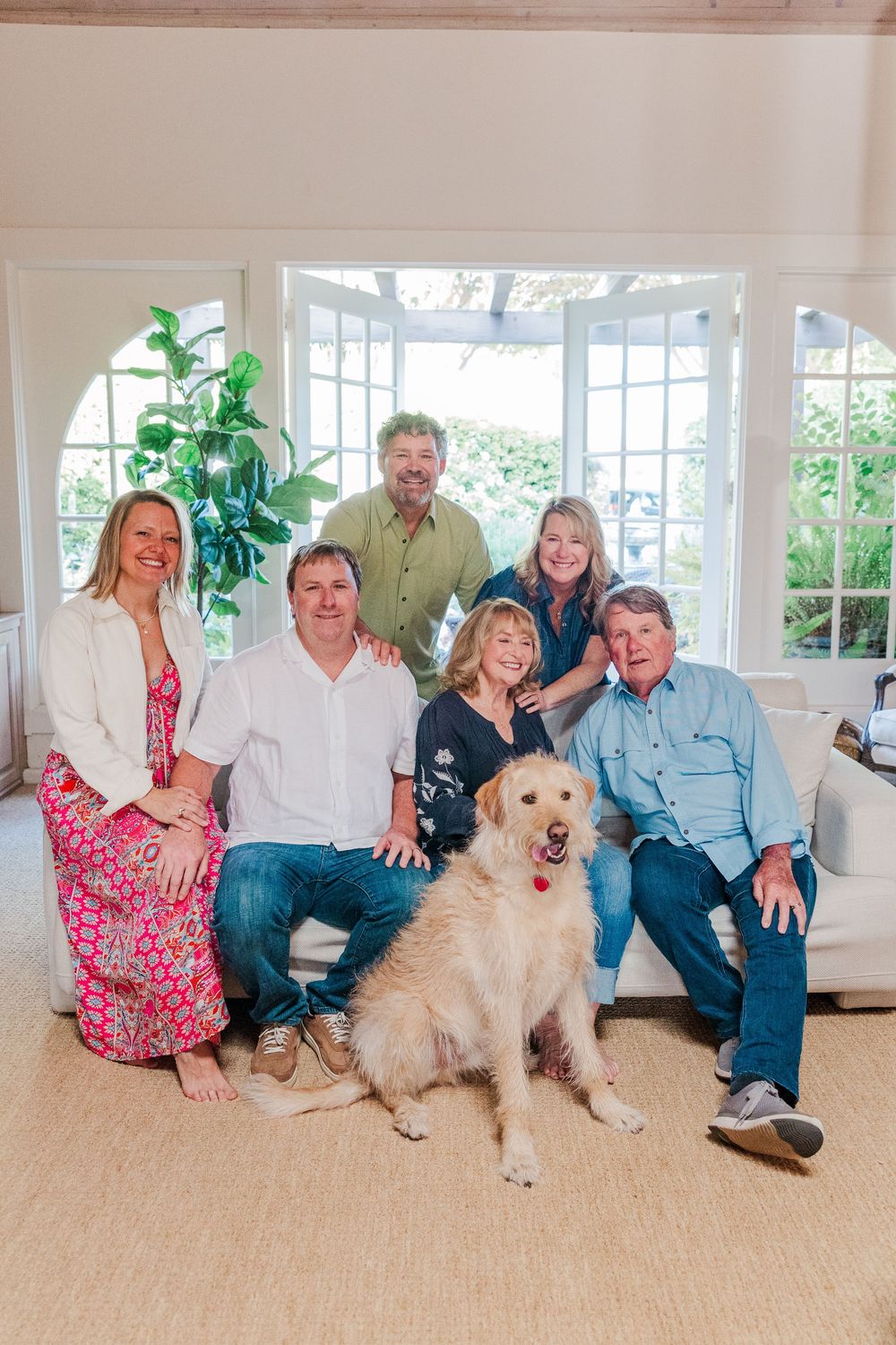 A group relaxes together on a bright sunlit couch with two fluffy white dogs in a coastal-style living room.