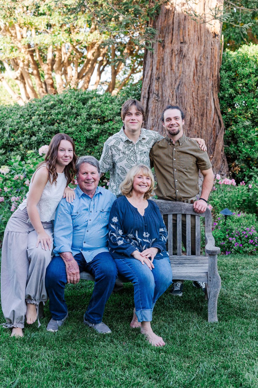 A group sits together on a wooden bench in a garden setting with a large tree trunk visible in the background.