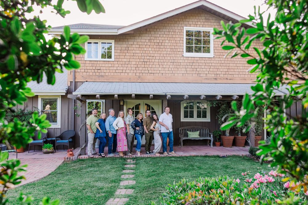 A residential house with tan siding and green landscaping features a group of people gathered near the entrance.
