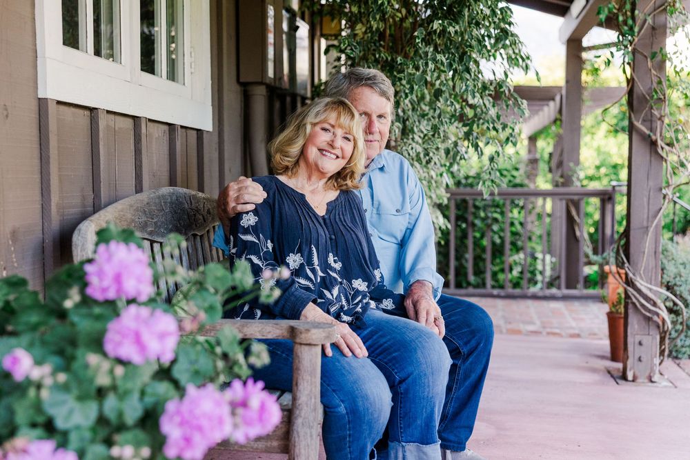 A couple sits together on a porch swing with pink roses blooming in the foreground.