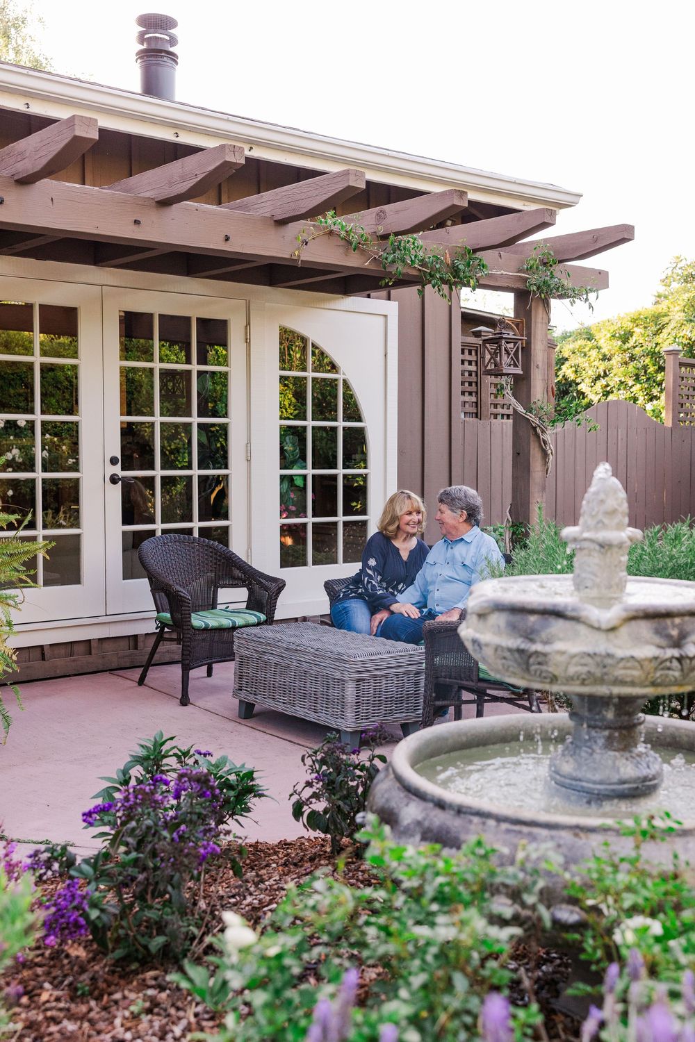 Elegant outdoor patio area with pergola, comfortable seating, and decorative stone fountain surrounded by purple flowers.