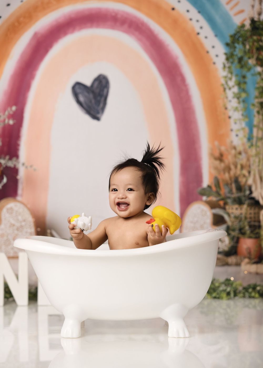 Smiling baby sits in vintage-style white bathtub against colorful rainbow backdrop in outdoor setting.