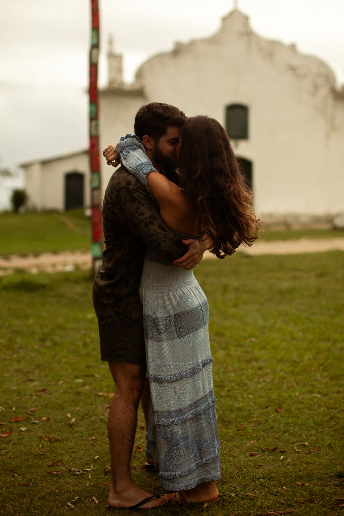 A couple embraces in a tender moment on a grassy field with a white building in the background.