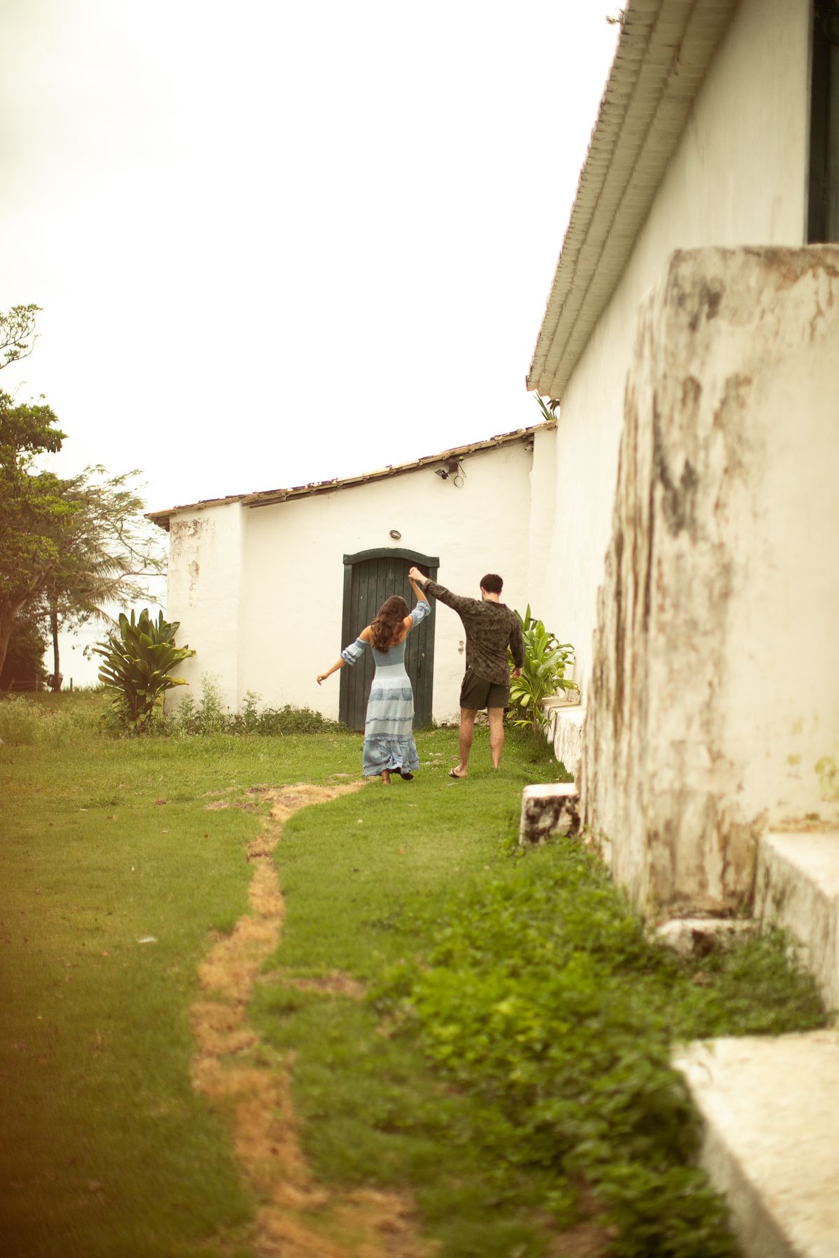 A dirt path leads to a white building entrance with people standing near the doorway in a rural setting.