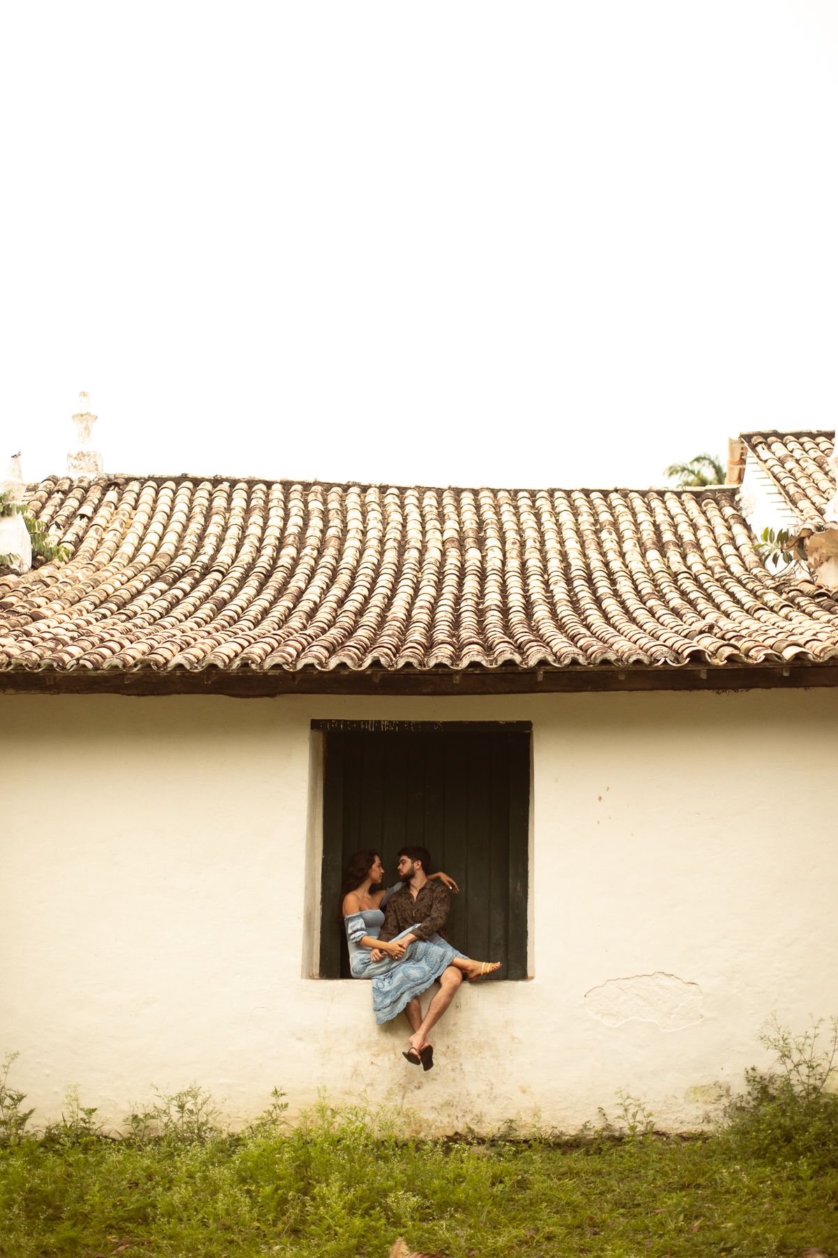 Two figures sit in a rustic window frame of a white building with traditional tiled roof.
