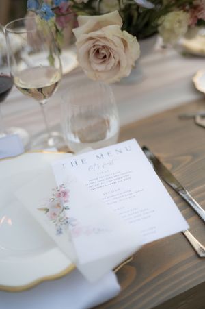 Wedding menu cards with floral watercolor designs sit on a rustic wooden table next to wine glasses and roses.