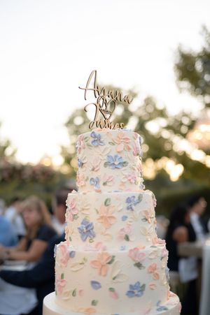 Three-tiered white wedding cake decorated with delicate pink and blue sugar flowers against an outdoor sunset backdrop.