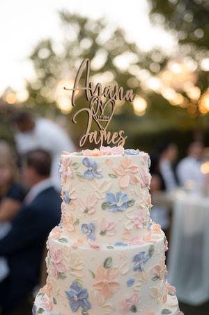 Elegant white wedding cake decorated with delicate pink and blue floral designs and a gold calligraphy topper at sunset.