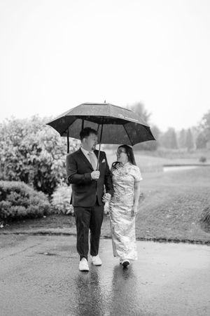 A couple shares an umbrella while walking on a rainy path in black and white wedding photography.
