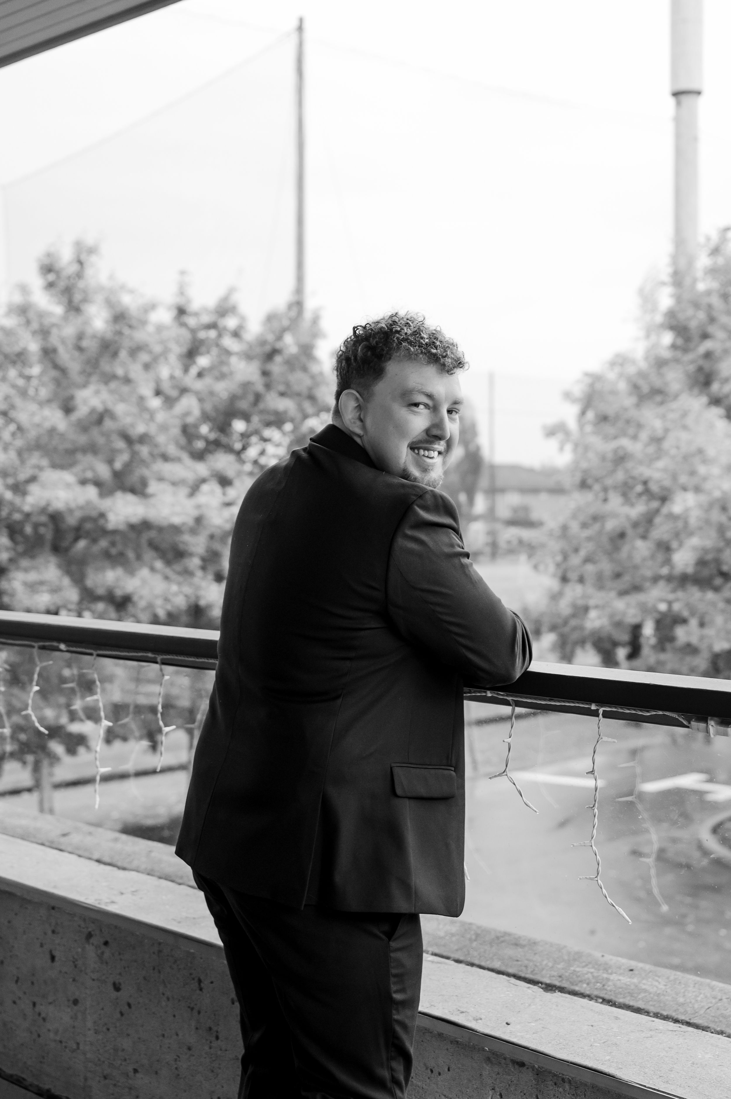 A black and white photo shows someone in a dark suit leaning on a railing outdoors.