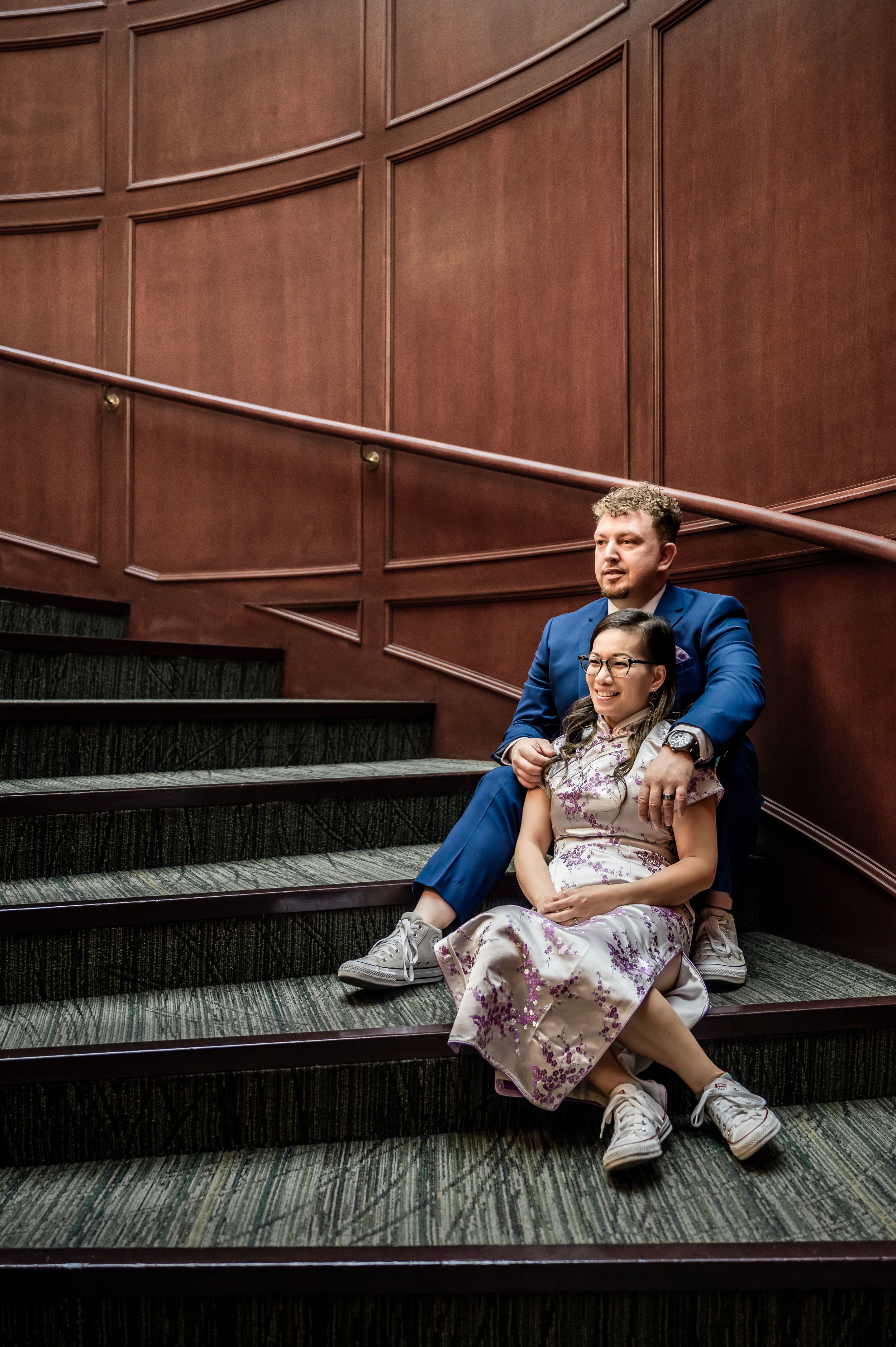 A person sits on wooden steps against a brown wall wearing a white dress and blue jacket.