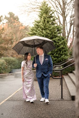 A couple in formal attire share black umbrellas while walking along a path surrounded by evergreen trees.
