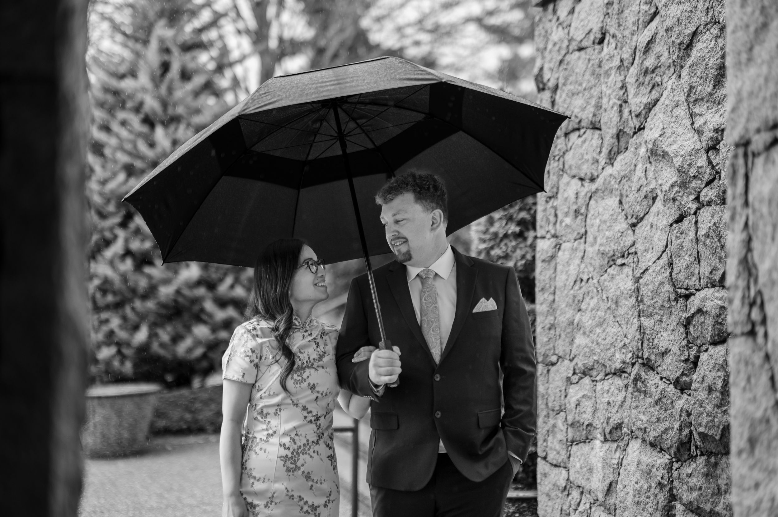 A couple shares a romantic moment under a black umbrella next to a stone wall in black and white photography.
