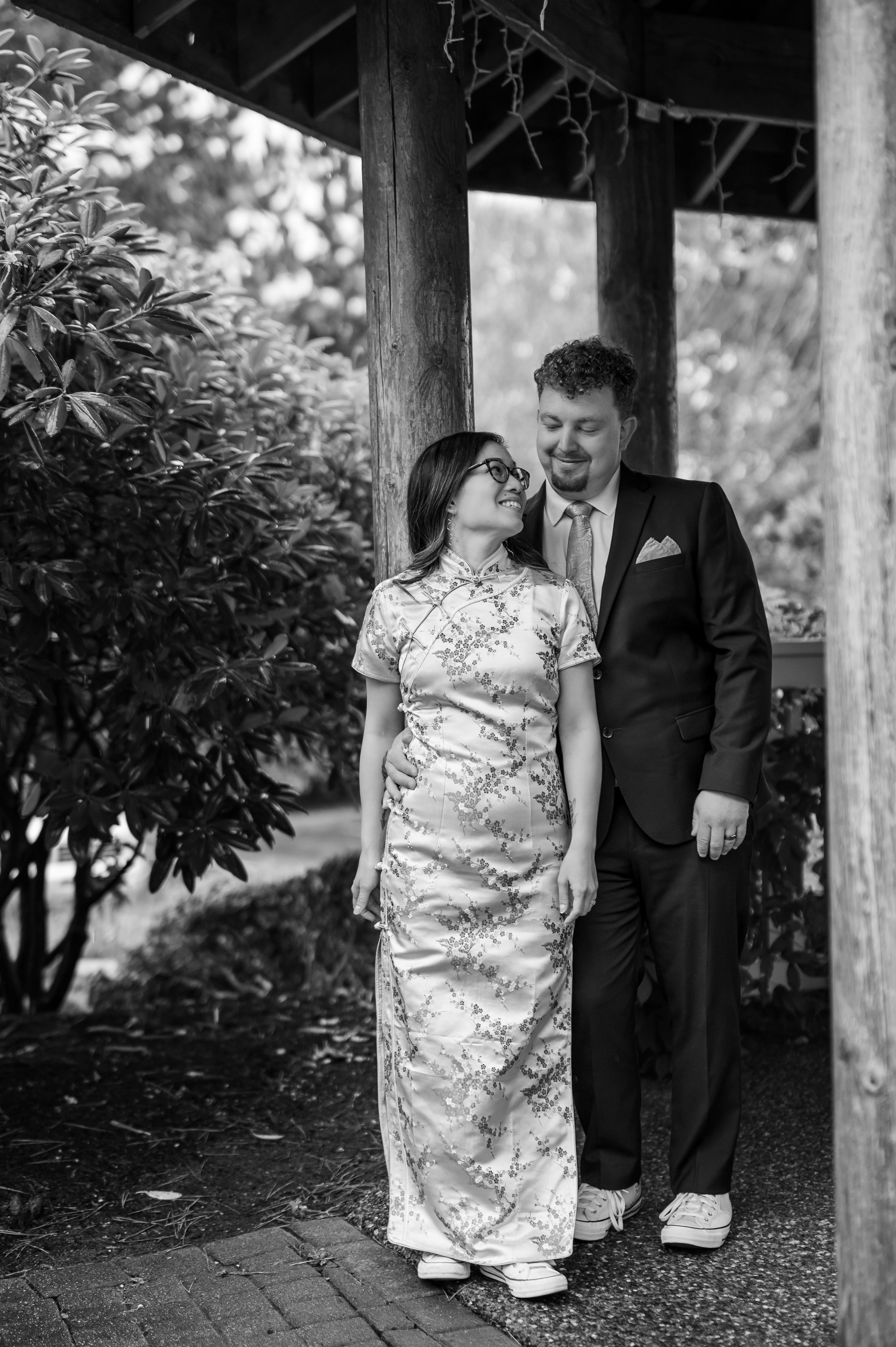 A couple in formal attire share a tender moment under a garden pergola in black and white photography.