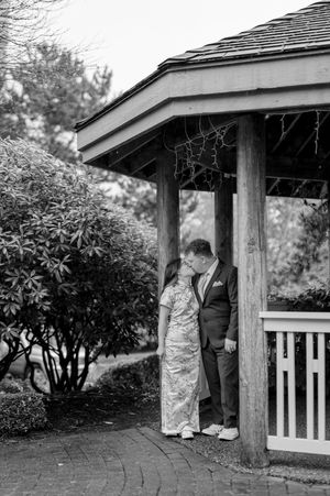 A couple stands together under a covered gazebo entrance in a garden setting, captured in black and white photography.