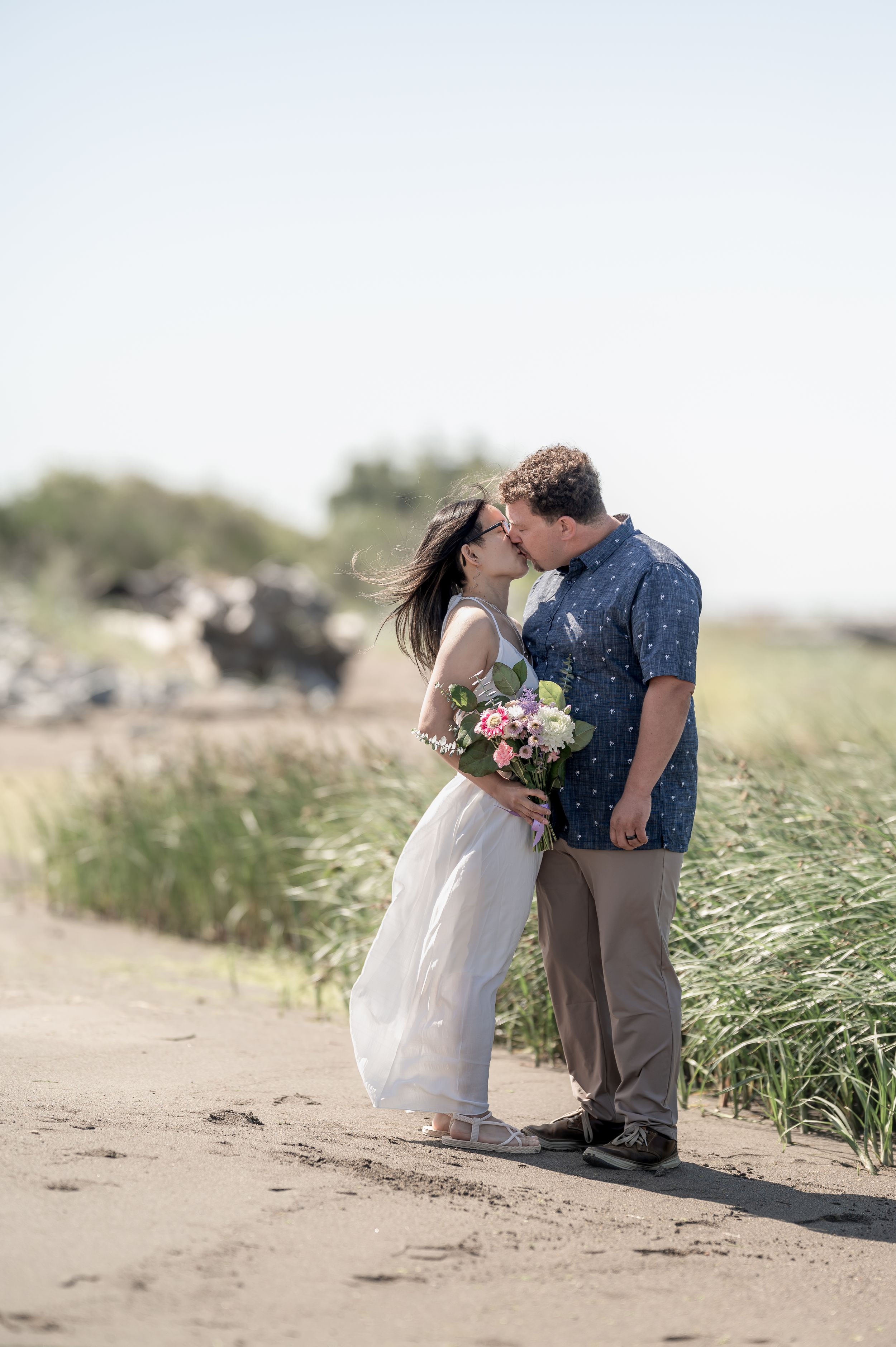A romantic couple shares a tender moment on a sandy beach path surrounded by windswept grass and holding a floral bouquet.