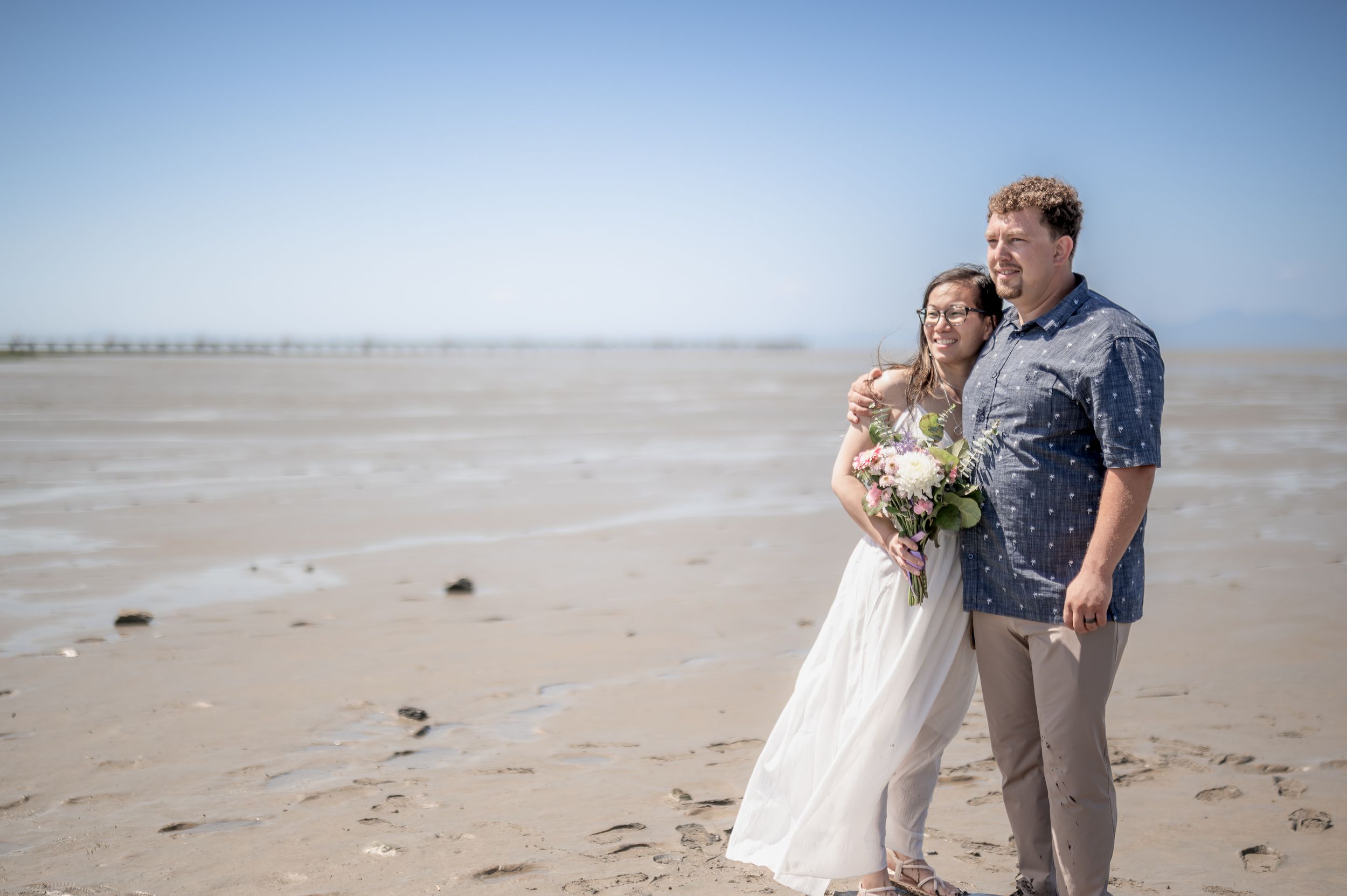 A couple shares a romantic moment on a sandy beach with the ocean and blue sky in the background.