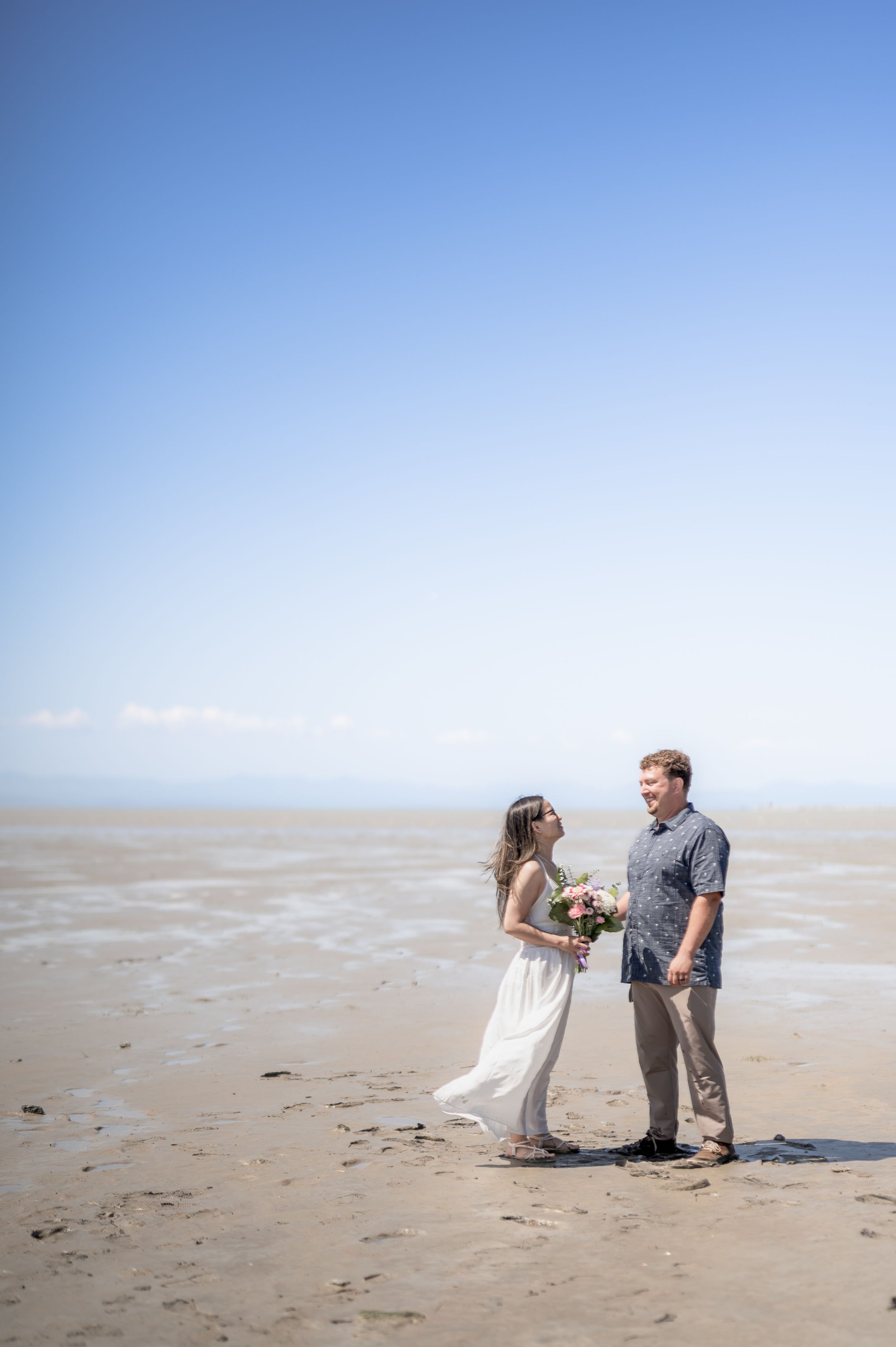 A couple shares tender moments on a sunny beach with a bright blue sky and sandy shore.